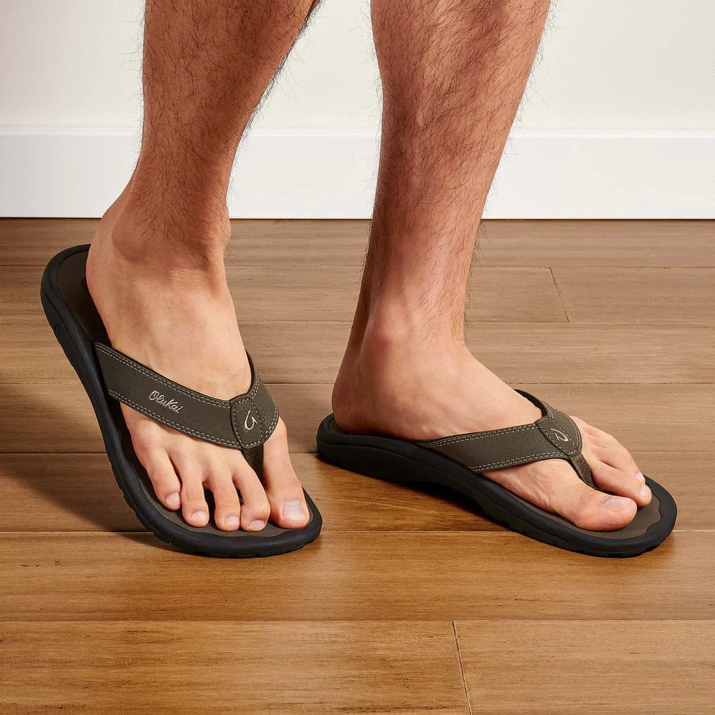 OluKai Ohana flip-flops in dark brown with water-resistant synthetic straps and cushioned footbed, worn indoors on wooden flooring.