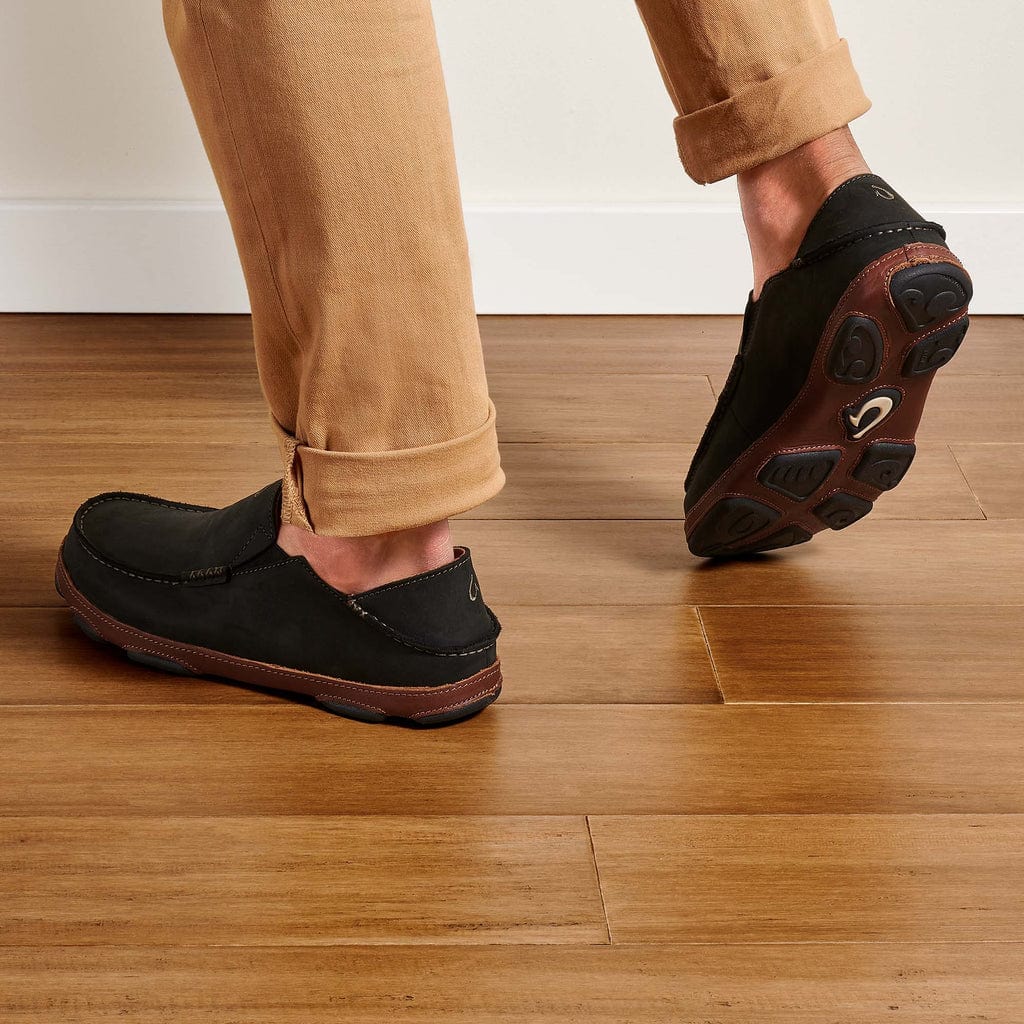 OluKai Moloa slip-on shoes in black with moccasin-style stitching and brown leather outsole, worn by man indoors.