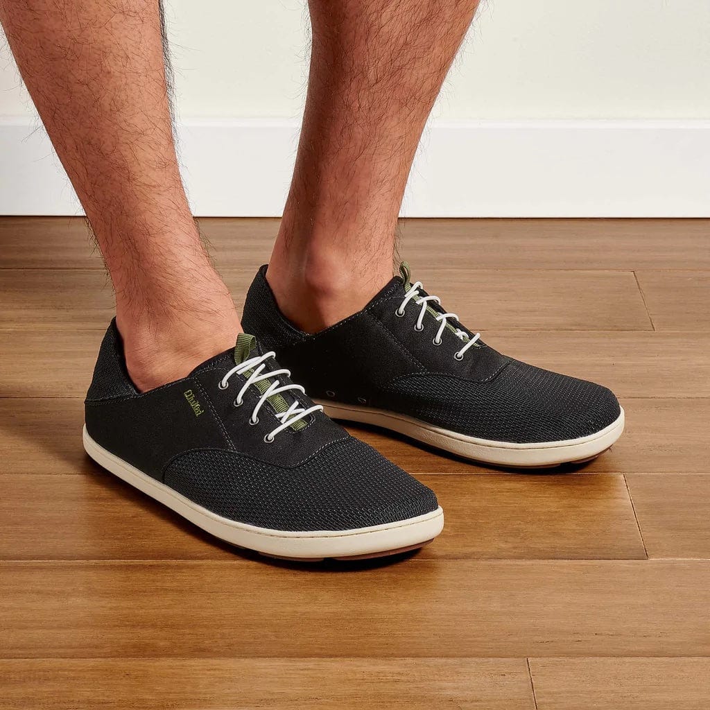 OluKai Nohea Moku casual shoes in black with breathable mesh upper, white laces, and lightweight beige sole, worn indoors.