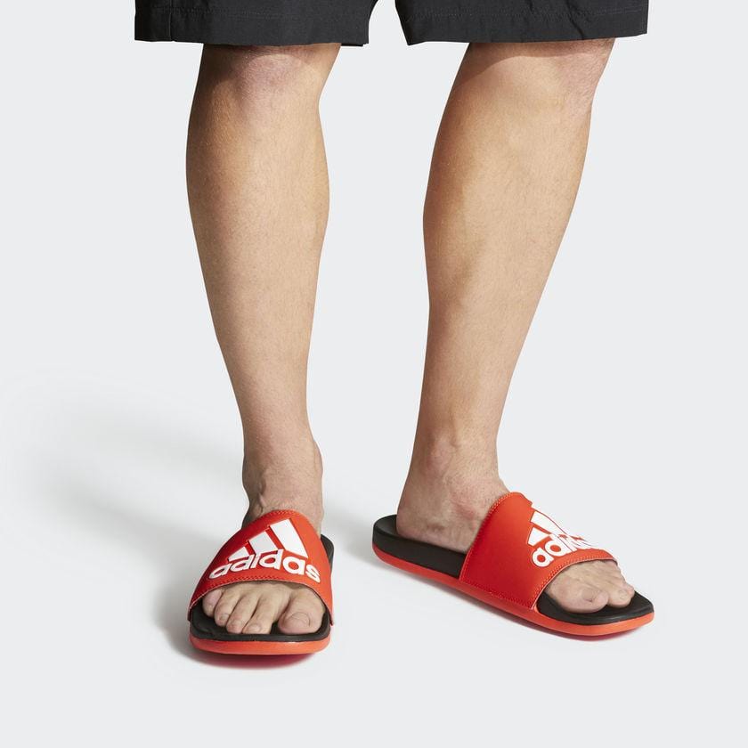 Adidas Adilette Comfort Slides in bold red with white Adidas branding on strap, cushioned black footbed, worn by person standing.