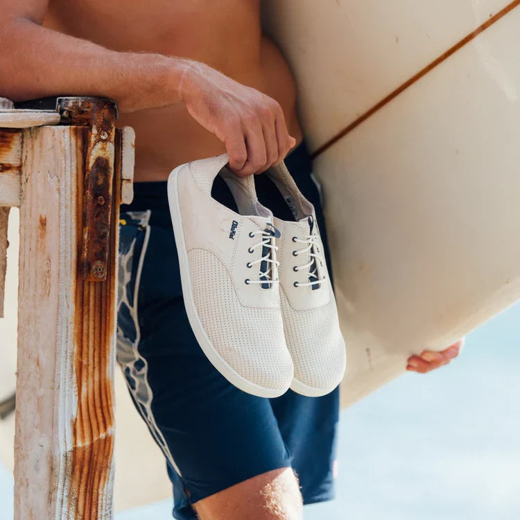 Person holding a surfboard and white sneakers with a beach setting