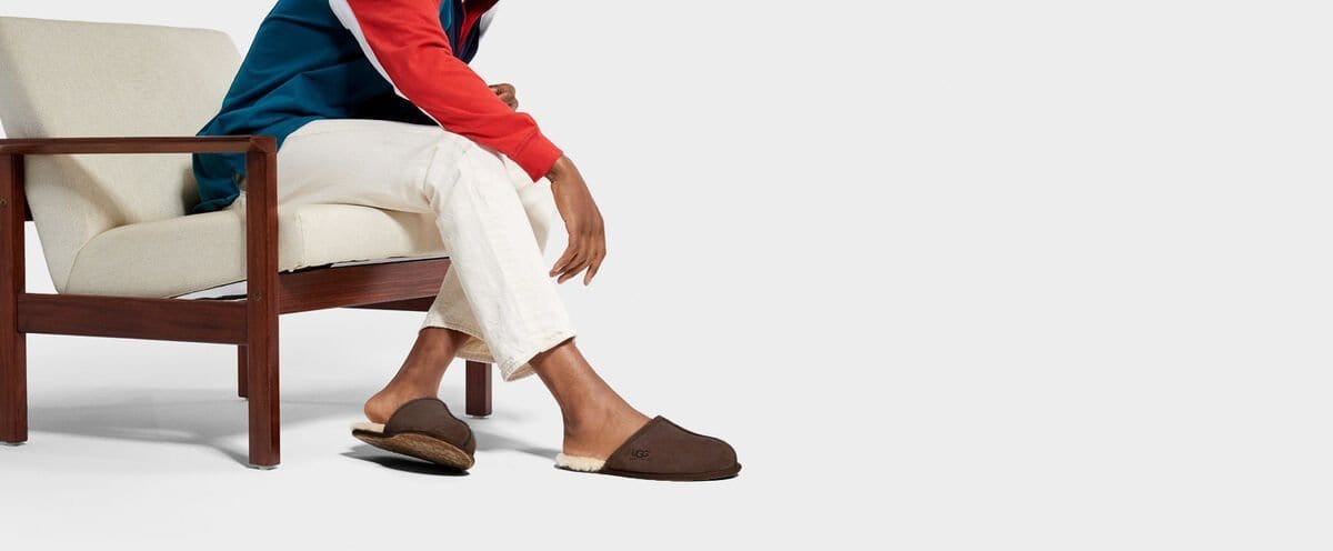 UGG Scuff slippers in dark brown suede with logo detailing and plush shearling lining, worn by seated person indoors.