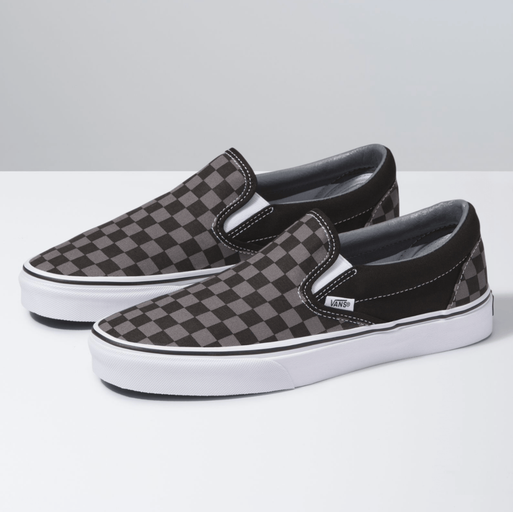 Vans Checkerboard Slip-On shoes in black and gray canvas with a classic checkered pattern, white midsoles, and elastic side accents, angled front side view.
