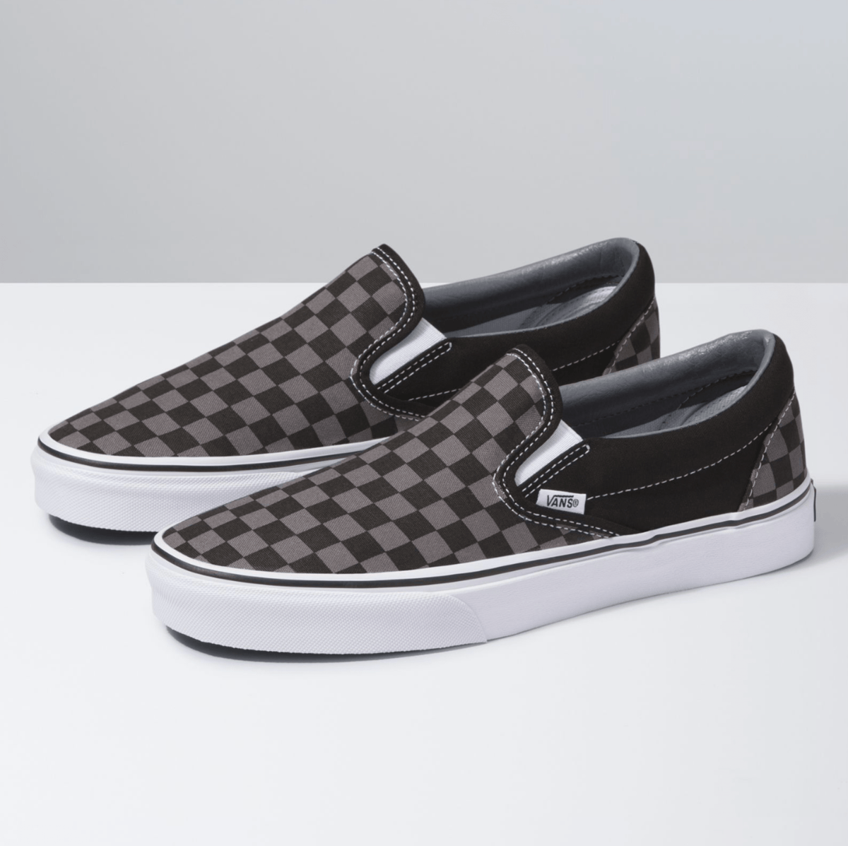 Vans Checkerboard Slip-On shoes in black and gray canvas with a classic checkered pattern, white midsoles, and elastic side accents, angled front side view.