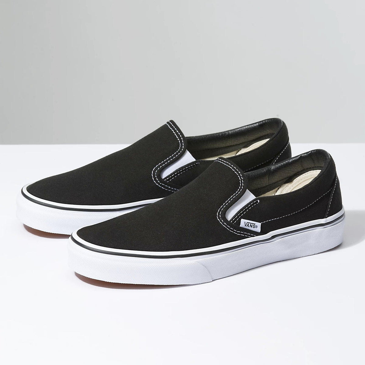 Vans Classic Slip On shoes in black canvas with white elastic side accents and signature white rubber sole, side angle view.