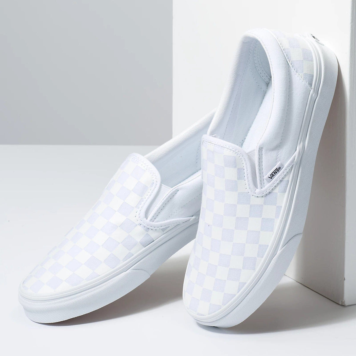 Vans Checkerboard Slip On shoes in white and light gray canvas with elastic side accents and waffle outsole, angled view.