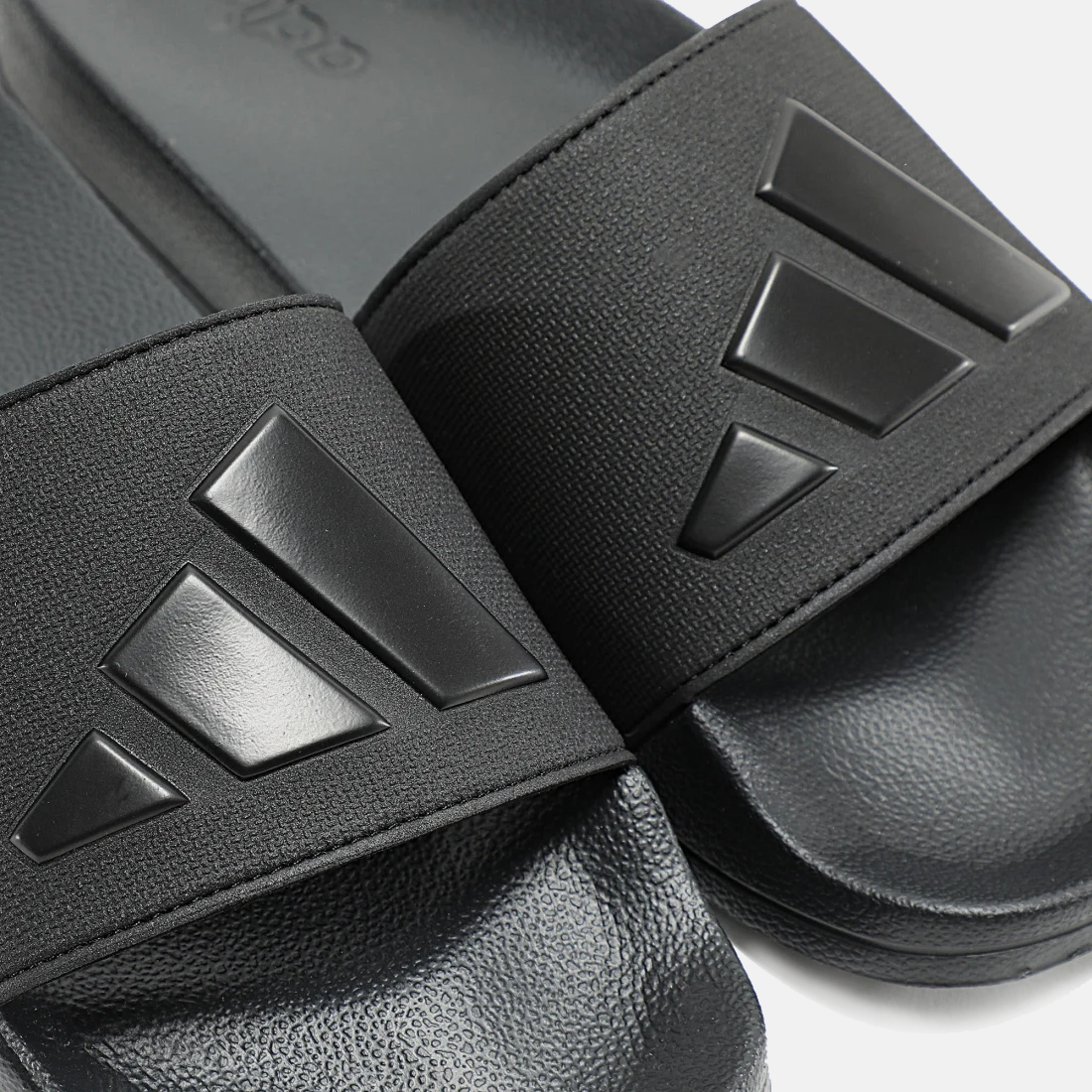 Adidas Adilette Shower Slides in black with textured footbed, synthetic strap, and embossed logo detail, close-up view.